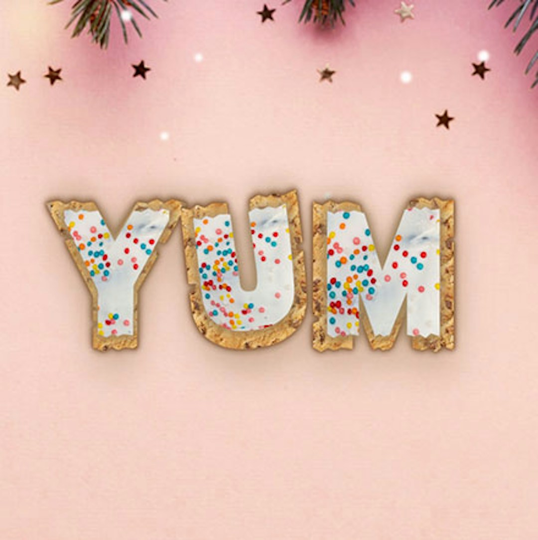 Text that says yum with a cookie base and white icing and sprinkles on top the background is a christmas theme